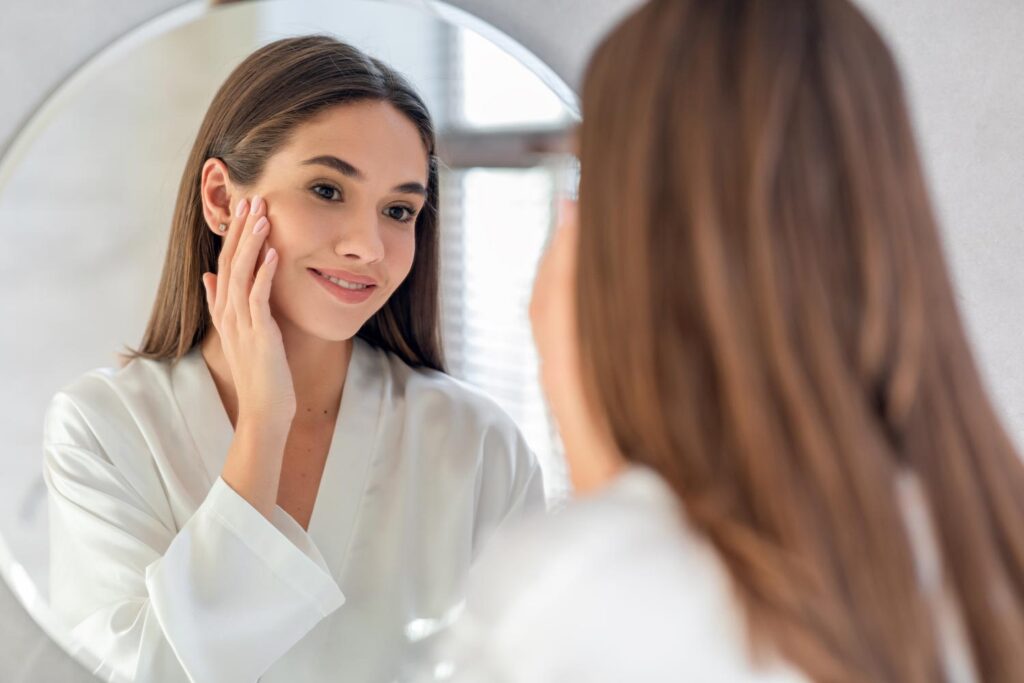 Woman smiling at her reflection in a round mirror, touching her cheek in a skincare routine