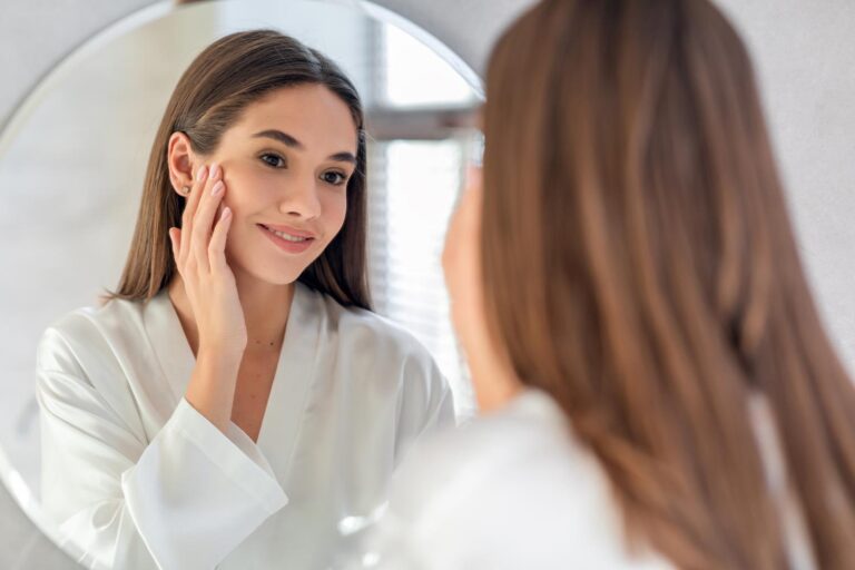 Woman smiling at her reflection in a round mirror, touching her cheek in a skincare routine