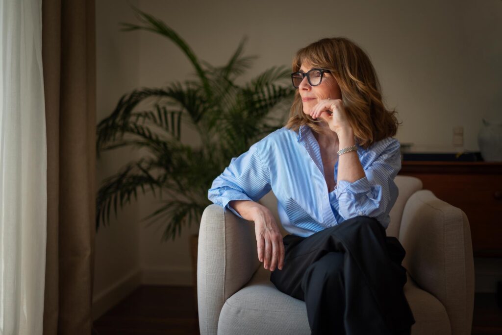 Woman with shoulder-length brown hair and glasses sits on a beige armchair by a window, resting her chin on her hand.