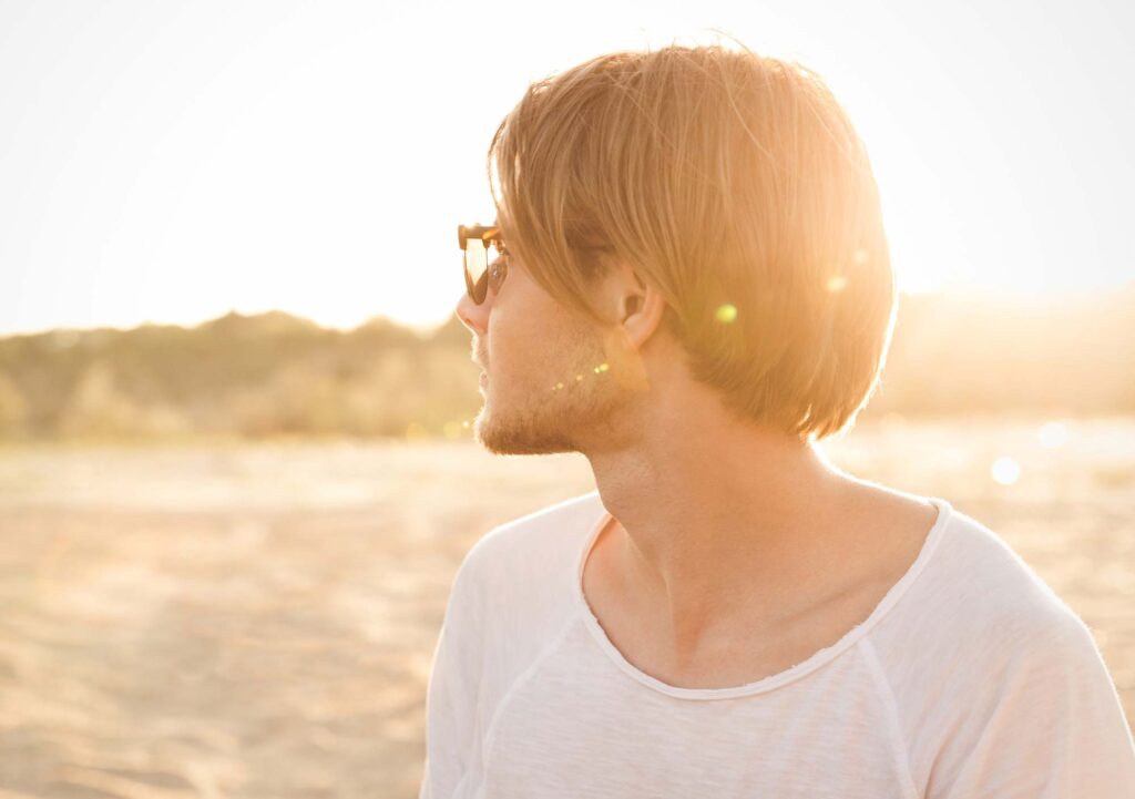 pcps-jaw-lift-neck-men - Pacific Center Plastic Surgery Side profile of a man with blond hair wearing sunglasses at the beach during sunset.
