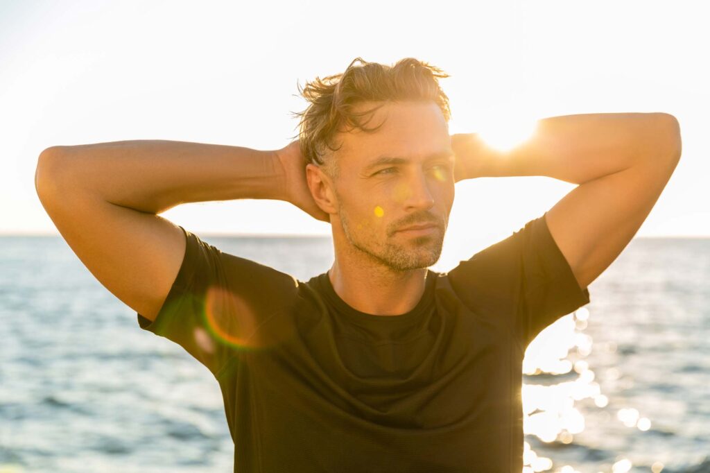 pcps-male-neck-lift - Pacific Center Plastic Surgery Man standing on a beach with hands behind his head, sun shining from behind him (backlit)