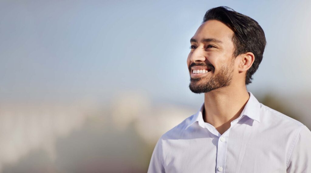 pcps-neck-jaw-lift-men - Pacific Center Plastic Surgery Smiling man with dark hair and beard wearing a white shirt outdoors against a blue sky backdrop