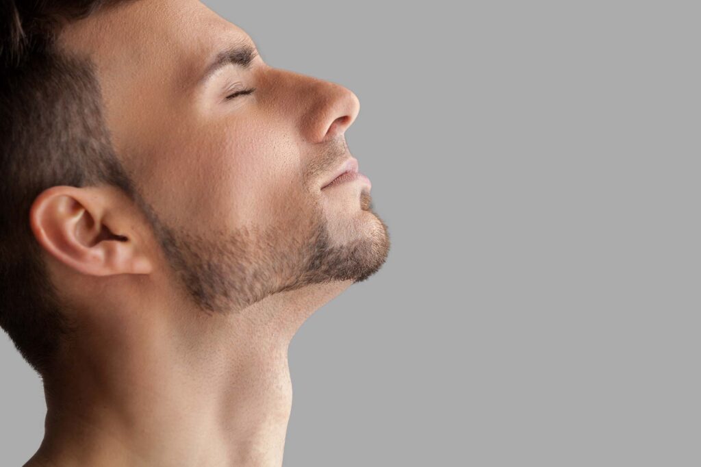 pcps-neck-lift-men - Pacific Center Plastic Surgery Close-up profile of a man with chin raised and eyes closed against a gray background.